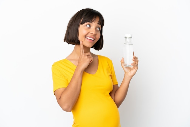 Jonge zwangere vrouw die een fles water vasthoudt die op een witte achtergrond wordt geïsoleerd en een idee denkt terwijl ze omhoog kijkt