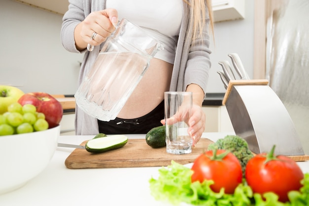 Jonge zwangere vrouw die water in glas op keuken giet