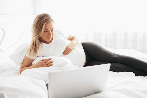 Jonge zwangere vrouw kijken naar film op laptop liggend op bed