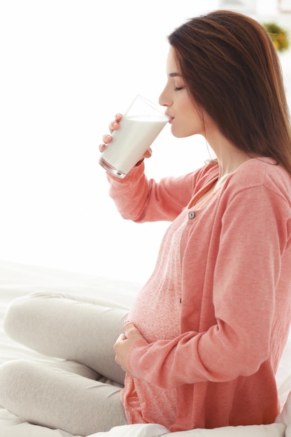 Jonge zwangere vrouw met glas melk zittend op bed in de kamer