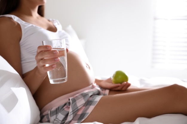 Jonge zwangere vrouw met glas water en appel in slaapkamerclose-up Het verzorgen van babygezondheid