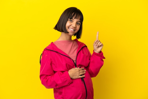 Jonge zwangere vrouw over geïsoleerde gele achtergrond die een vinger toont en opheft in teken van het beste