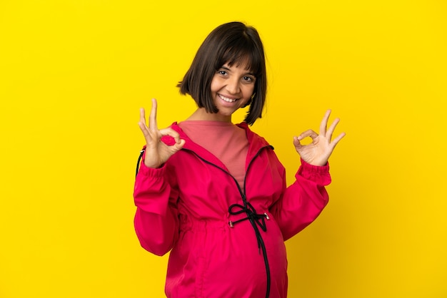 Jonge zwangere vrouw over geïsoleerde gele achtergrond die ok teken met twee handen toont