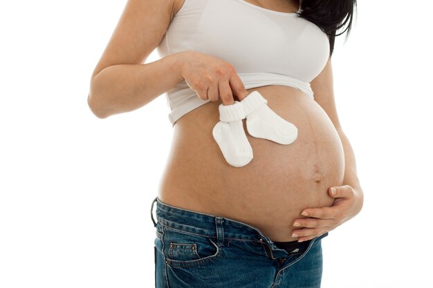 Jonge zwangere vrouw poseren in wit overhemd met kleine sokken in haar handen geïsoleerd op de muur