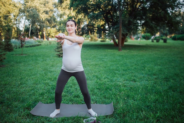 Jonge zwangere vrouwentribune op yogapartner buiten in park. Ze strekt haar armen uit en kijkt vooruit. Model ziet er geconcentreerd uit.