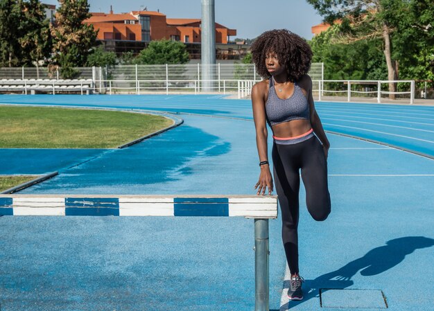 Jonge zwarte atleet meisje met afro haar dat zich uitstrekt staande leunend op een houten hek, gekleed in zwarte sportkleding en een grijze top met blauwe atletiekbaan op de achtergrond