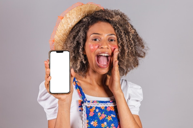Jonge zwarte Braziliaanse vrouw met landelijke kleding gekleed voor Festa Junina met smartphone met wit scherm voor advertenties mobiele apps