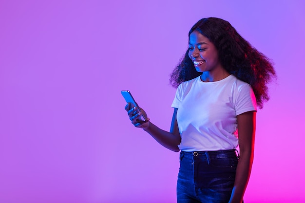 Jonge zwarte dame die mobiel surft op internet en geniet van moderne technologieën die nieuwe mobiele app in neonlicht controleren