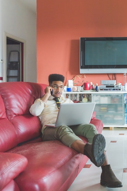 Jonge zwarte man binnenzittinglaag die laptop computer met behulp van die slimme telefoon spreekt