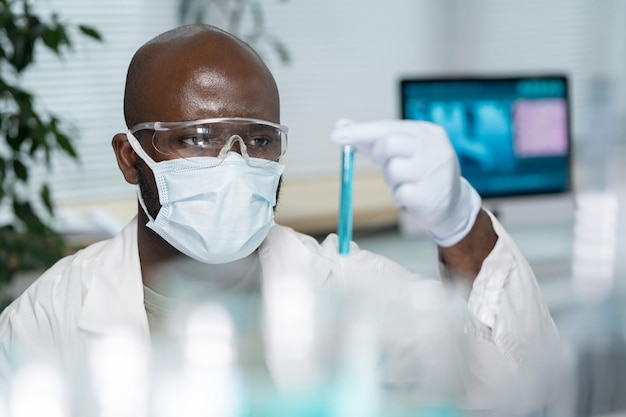 Jonge zwarte man in laboratoriumjas beschermend masker en handschoenen kijken naar kolf