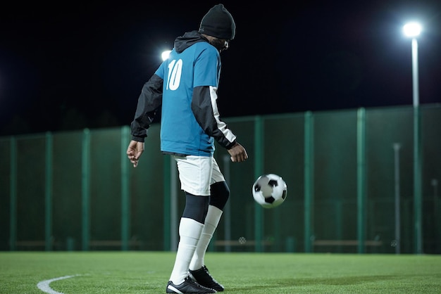 Foto jonge zwarte man in professioneel voetbaluniform die voetbal schopt tijdens het spelen op het veld omringd met verlichting