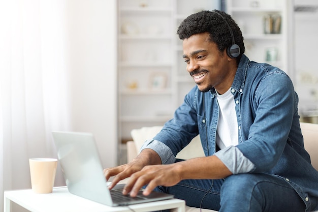 Jonge zwarte man met koptelefoon die binnen op een laptop werkt