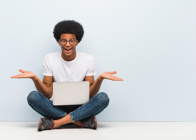 Jonge zwarte man zittend op de vloer met een laptop die twijfelt en schouders ophaalt