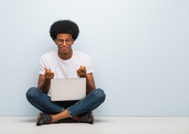 Jonge zwarte man zittend op de vloer met een laptop doet een gebaar van noodzaak