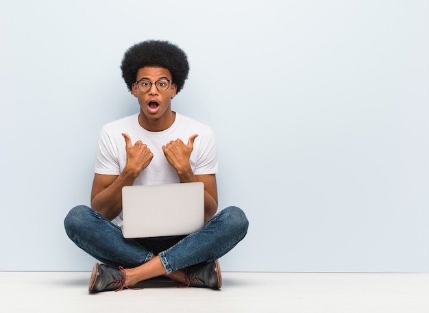 Jonge zwarte man zittend op de vloer met een laptop verrast, voelt zich succesvol en welvarend