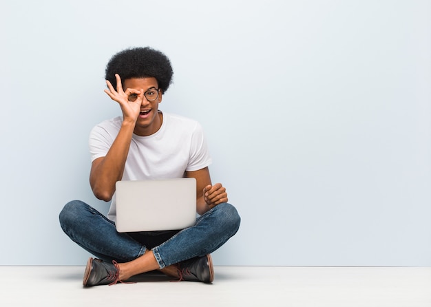 Jonge zwarte man zittend op de vloer met een laptop zelfverzekerd doen ok gebaar op oog