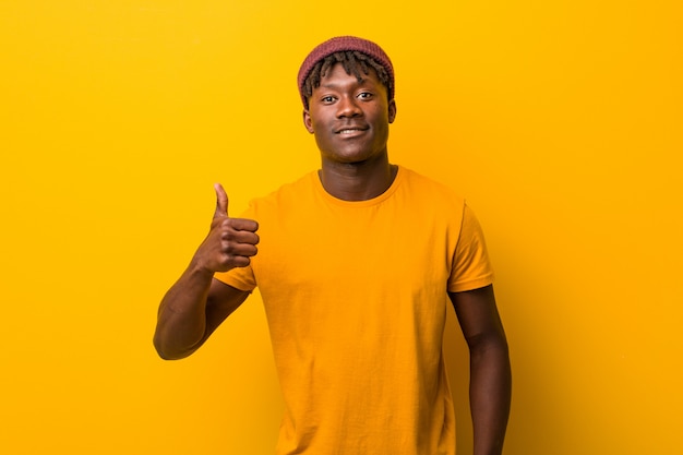 Jonge zwarte mens die rastas over gele achtergrond draagt die en duim glimlacht opheft