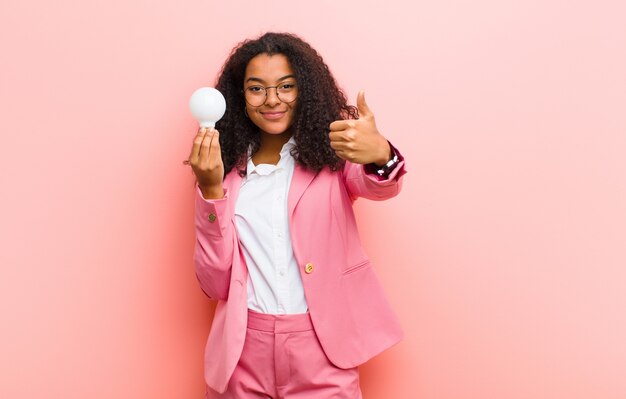 Jonge zwarte mooie vrouw met een gloeilamp die een idee heeft tegen roze muur