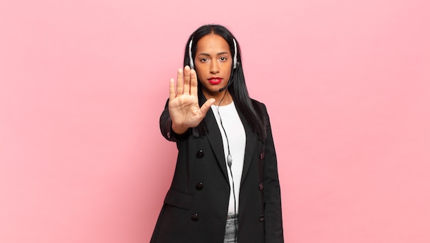 Jonge zwarte vrouw die er serieus, streng, ontevreden en boos uitziet met een open palm die een stopgebaar maakt. telemarketing concept
