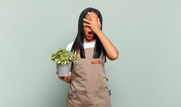 Jonge zwarte vrouw die geschokt, bang of doodsbang kijkt, haar gezicht bedekt met de hand en tussen de vingers gluurt. tuinman concept