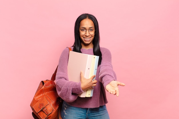 Jonge zwarte vrouw die vrolijk lacht met een vriendelijke, zelfverzekerde, positieve blik, een object of concept aanbiedt en toont. studentenconcept