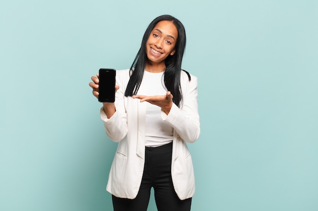 Jonge zwarte vrouw lacht vrolijk, voelt zich gelukkig en toont een concept in kopieerruimte met de palm van de hand. slimme telefoon concept