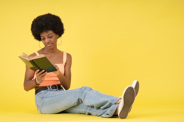 Jonge zwarte vrouw met afrokapsel zittend op de vloer en zich concentrerend op het lezen van een boek