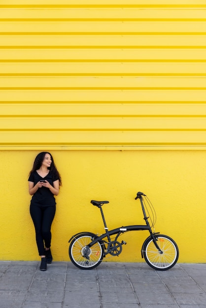 Jonge zwarte vrouw met lang haar en zwarte outfit sms't iemand naast haar fiets voor een gele muur