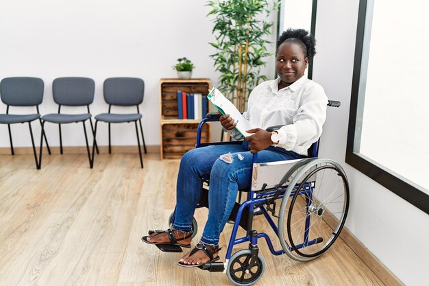 Jonge zwarte vrouw zittend op een rolstoel in de wachtkamer glimlachend naar de zijkant kijkend en weg starend denkend