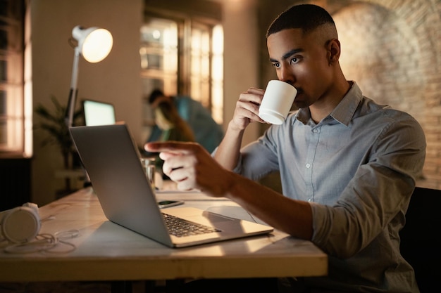 Jonge zwarte zakenman die op een computer werkt en 's nachts koffie drinkt op kantoor