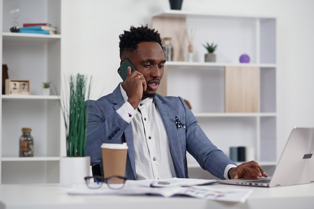 Jonge zwarte zakenman die op mobiele telefoon spreekt en aan laptop in modern wit bureau werkt, exemplaarruimte