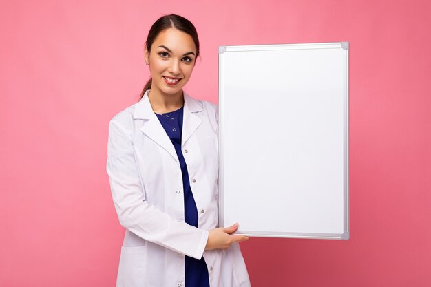 Jongelui van mooie gelukkige donkerbruine vrouw die medische witte jas draagt die wit magnetisch bord houdt dat op roze muur met lege ruimte wordt geïsoleerd.