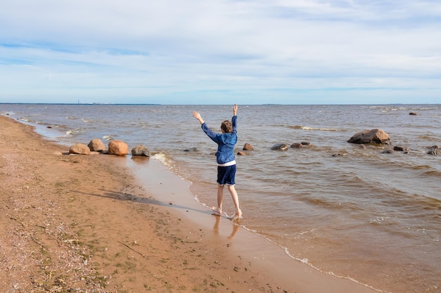 Jongen blootsvoets rennen op zand met handen vrolijk omhoog, vanaf de achterkant aan de kust in blauwe jas en korte broek, zonnige dag