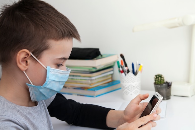 Jongen die thuis met een smartphone in zijn handen en een medisch masker onderwijst. Coronavirus Quarantaine Concept