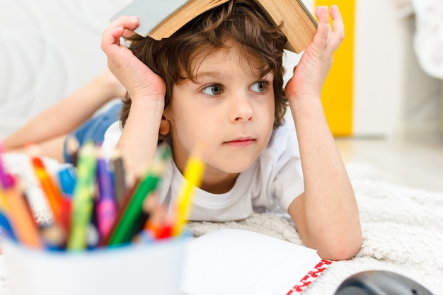 Jongen doet huiswerk en houdt een boek op zijn hoofd in lichte kamer. Conceptlezing, onderwijs, jeugd. Sociale afstand nemen en zelfisolatie bij het afsluiten van quarantaine.