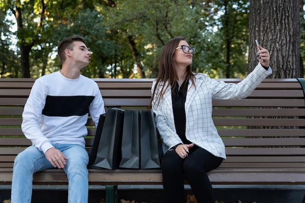 Jongen en meisje nemen selfie op bankje met boodschappentassen. Jonge man en vrouw rusten na het winkelen op een bankje in het park.