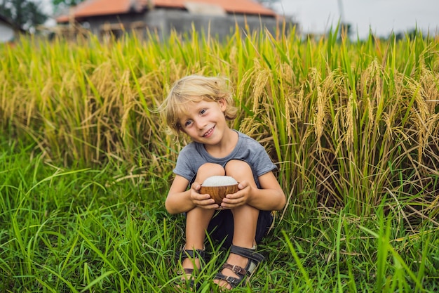 Jongen houdt een kopje gekookte rijst in een houten kopje op de achtergrond van een rijp rijstveld. Voedsel voor kinderen concept.