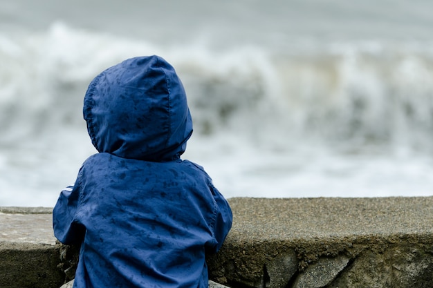 Jongen in blauwe jas met capuchon staat met zijn rug. Pier tegen de achtergrond van zee golven