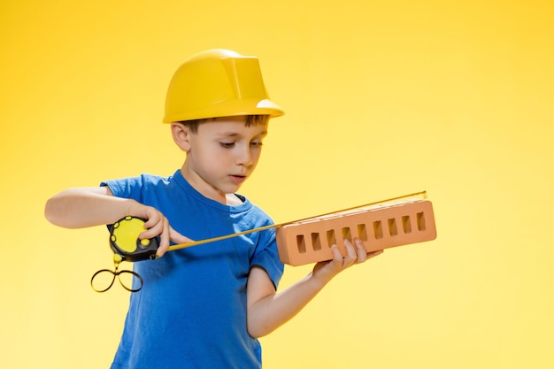 Jongen in een bouwhelm houdt een baksteen in zijn handen en meet deze met een meetlint in studio op gele achtergrond