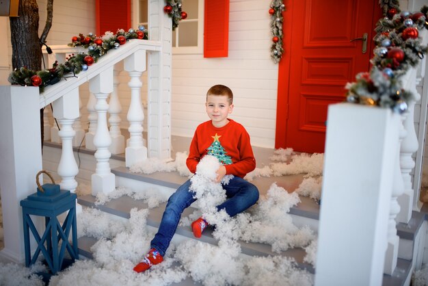 Jongen in een kersttrui zit op de trappen van een huis