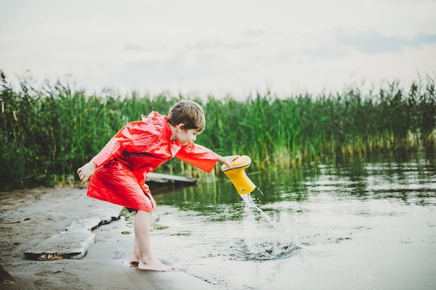 Jongen in een rode regenjas giet water uit gele rubberen laars in het meer, kind speelt met water