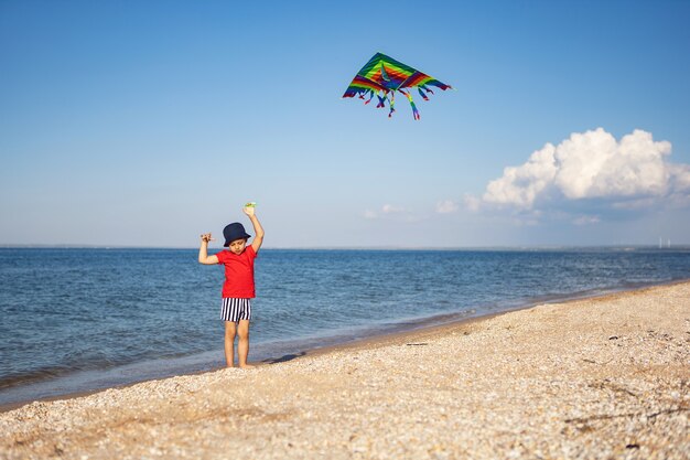 Jongen in een rood T-shirt en gestreepte zwembroek lanceert een vlieger op het strand aan zee in de zomer op vakantie