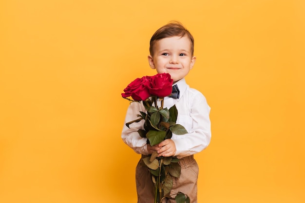 Jongen in een witte overhemdbroek en een vlinderdas op een gele achtergrond houdt in zijn handen een boeket rode rozen Een cadeau voor mijn moedergrootmoeder