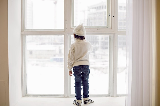 Jongen in een witte trui en hoed staat in de winter bij het raam in een groot appartement op eerste kerstdag