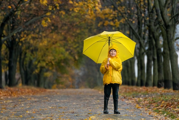 Jongen in gele regenjas met grote gele paraplu loopt door het herfstpark Gelukkig kind