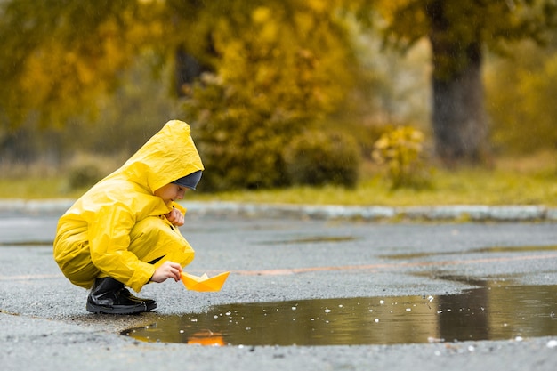 Jongen in gele waterdichte mantel en zwarte rubberen laarzen spelen met papieren handgemaakte boot speelgoed in een plas buiten in de regen in de herfst.