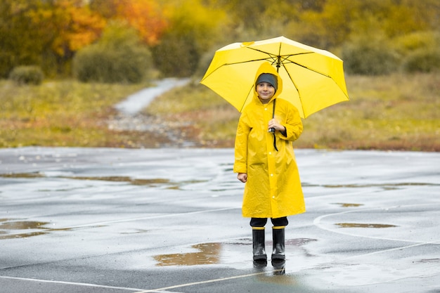 Jongen in gele waterdichte mantel en zwarte rubberen laarzen staan in een plas buiten in de regen in de herfst.