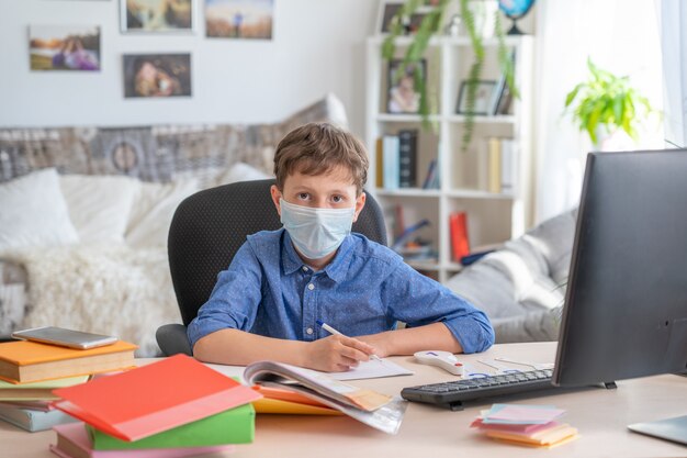 Jongen in gezichtsmasker met behulp van computer, huiswerk tijdens coronavirus quarantaine