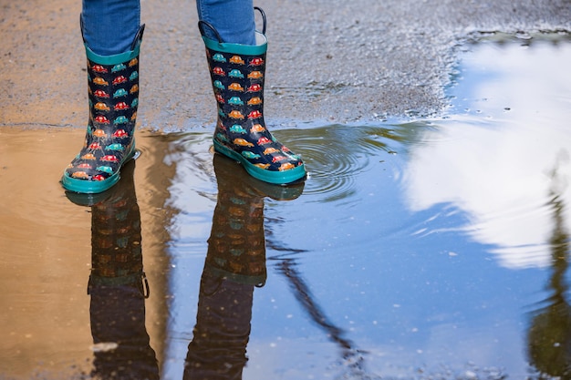 Jongen in grappige rubberen laarzen die na regen in de plas op straat staat Paar kleurrijke rubberen laarzen in een plas