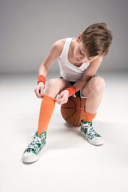 Jongen in sportkleding zittend op basketbal bal geïsoleerd op grijs